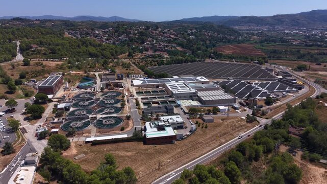 Large wastewater treatment plant with circular clarifier tanks and solar panels, surrounded by green hills and a residential town in a Mediterranean landscape