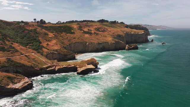 Rows of white surf roll toward sandstone headlands and steep cliffs at Tunnel Beach. An aerial pass reveals golden slopes and a coastal walking path near Dunedin, New Zealand.