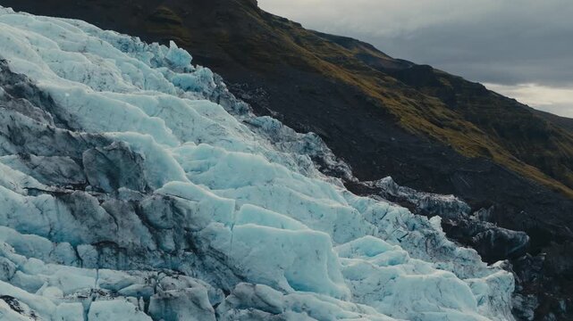 Steep fractured blue glacier ice meets dark rocky terrain in Iceland, revealing dramatic layers, crevasses and rugged glacial geology - drone shot