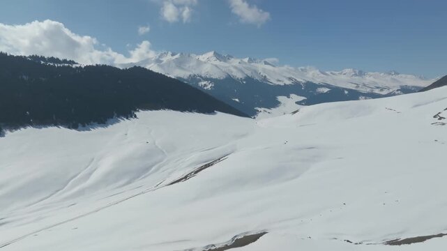 Panoramic footage of the vast snow covered Grigorievskoye gorge valley in Issyk-Kul Region, Kyrgyzstan, showing evergreen pine forests and jagged majestic mountain peaks under blue sky