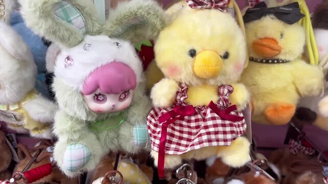 Cute yellow duck and anime doll stuffed animal keychains. Adorable fluffy chick keyring dressed in a red plaid checkered dress alongside a green rabbit ear doll on retail gift shop accessory displays.