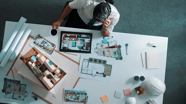 Top view of engineer drink coffee and thinking about house plan at meeting table with equipment and safety helmet scatter around. Aerial view of stressful manager looking at blueprint. Alimentation.