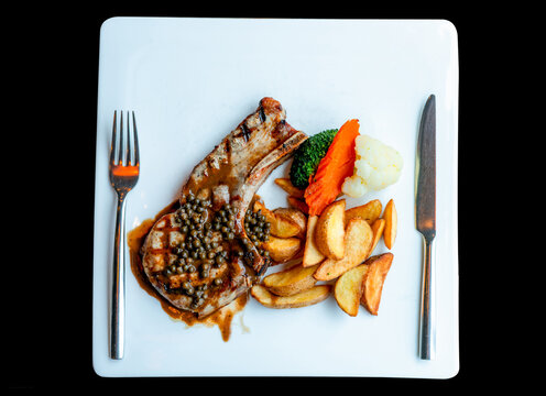 Grilled bone in meat chop topped with green peppercorns and brown sauce, served with golden potato wedges, broccoli, carrot and cauliflower on a white rectangular plate with a metal fork and knife.
