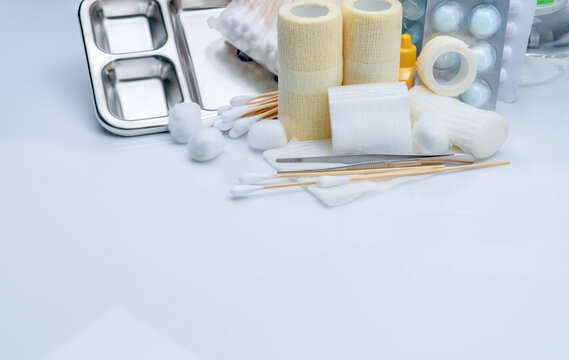 Wound care supplies on a white surface, rolled bandages and folded gauze beside cotton swabs, metal forceps, a stainless steel tray and bottled liquids, a neat arrangement for first aid preparation