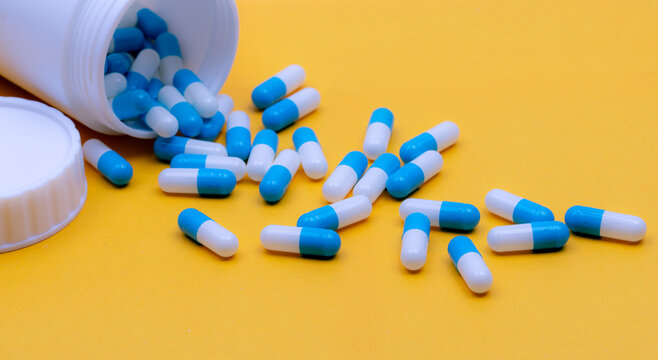 Blue and white prescription capsules spilling from an open white plastic bottle onto a yellow surface, with the detached screw cap beside the opening and more capsules remaining inside.