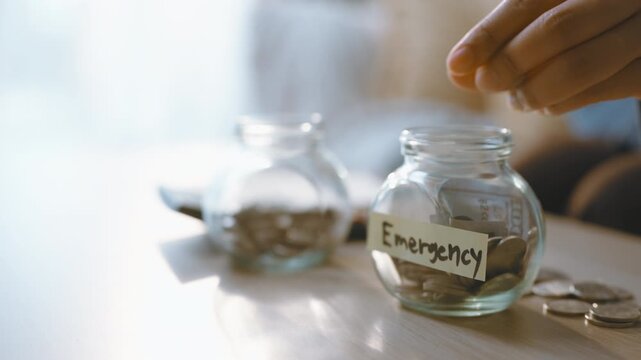 Emergency saving is represented by a hand dropping a coin into a labeled glass jar, with money nearby on the table as finance planning and saving for a reserve fund become a household concept.