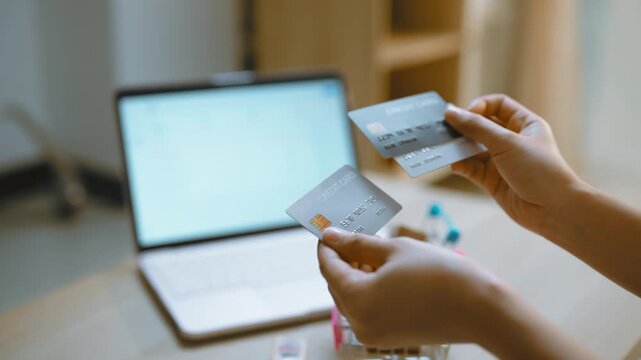 Payment card comparison beside a laptop symbolizes shopping with credit and debit options, as hands hold two cards near a miniature cart to represent a digital payment choice at a home table.