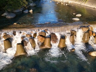 A close angled overhead image shows a line of small cascades breaking between large tan blocks into green blue water. © Masakazu Tokashiki