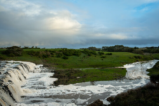 Powerful waterfall, Hopkins Falls, cascades over rocky escarpment into winding river, Hopkins River, surrounded by green rolling hills under expansive, storm-filled sky, Cudgee, Victoria, Australia
