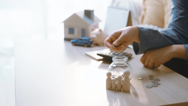 Saving money is represented by a man placing coins into a glass jar beside a calculator, with a model house and toy car nearby as finance planning and a household budget support the saving activity.
