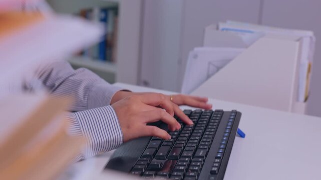 Person Typing on Computer Keyboard at Office Desk, Hand Working with Paperwork and File Organizer, Close Up of Focused Administrative Work in Quiet Workplace