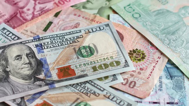 American 100 dollar (USD) bills falling on top of Thailand national currency (THB). Paper cash lands across colorful Thai baht, illustrating overseas remittances, global trade and personal wealth