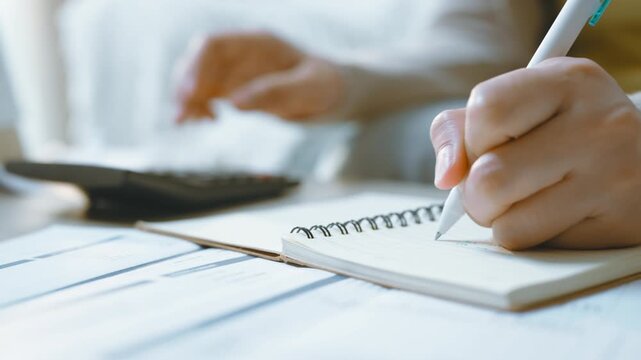 Writing in a notebook while using a calculator illustrates finance and accounting, with a pen moving across the page beside printed budget forms as hands organize a personal finance calculation.