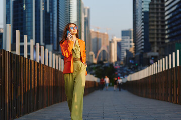 Obraz na płótnie Canvas Strategy consultant, confident young woman in orange blazer and sunglasses talking on smartphone walking across city bridge with skyscrapers. Concept of business success, urban lifestyle and ambition.