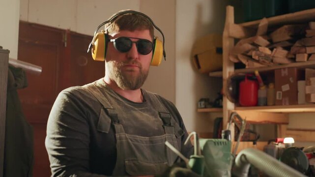 Man operating woodworking equipment with ear protection. Workshop environment includes shelves lighting machinery woodworker power tool sawdust tools gloves