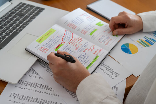 Hands writing goals and appointments in a weekly planner on a desk with laptop, financial reports and charts, depicting business planning, time management and organized work routine