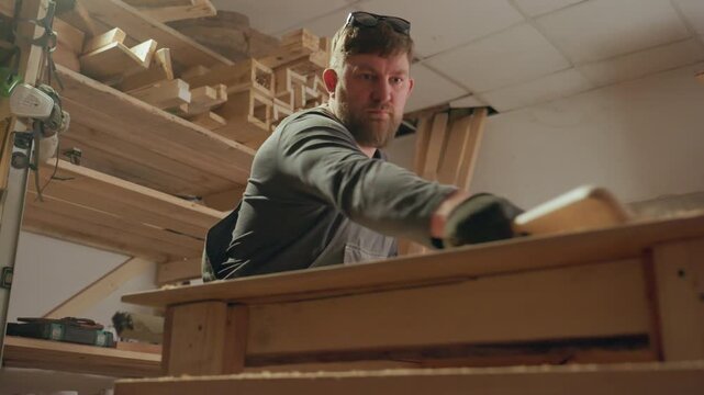 Man works at workbench shaping wood with various power and manual joinery tools. background shows shelves filled with lumber and workshop equipment under warm lighting