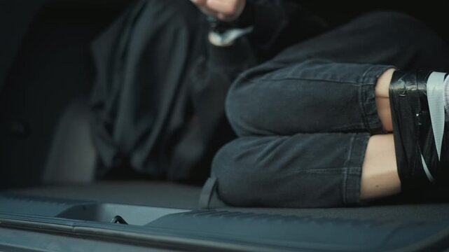 Woman lies restrained inside what appears to be car trunk. Her legs are tied together and her hands rest on edge of seat bound vehicle interior restraint