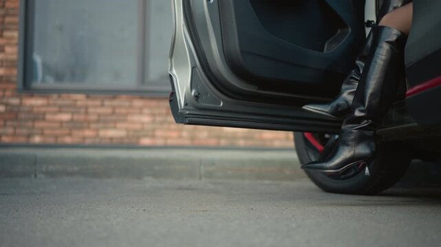 Shows woman black leather high heeled boots with pointed toes as woman steps out of open car door. scene includes lower part of her legs concrete ground and background brick wall