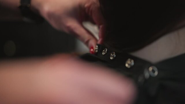 Close up shows skilled hand movements as hairstylist attaches extension focusing on technique and interaction with client natural hair. Hands manipulate black strap through metal grommets