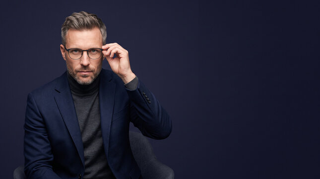 A man with grey hair wearing glasses, a dark blazer, and a grey turtleneck sweater, sitting ad social media download thambnail poster web template event festival 2026 2027 2028
