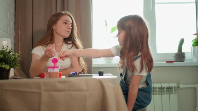 Woman sits at table engaged in art project while young girl reaches out to assist or interact with painting process. scene is set indoors near natural light featuring household items