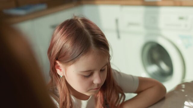 Girl rests her arms on countertop inside bright domestic setting. Girl has light brownish red hair and wears small earrings. washing machine is visible blurred in background suggesting