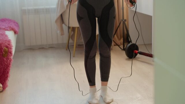 Woman in white sports bra and gray patterned leggings prepares with her hands on handle of electronic jump rope. Woman stands indoors near bright windows surrounded by home decor