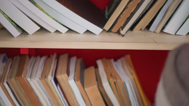 Shows red wooden shelf filled with various bound volumes. human finger reaches towards bottom row of older hardcover texts. Soft natural light illuminates scene highlighting book spines