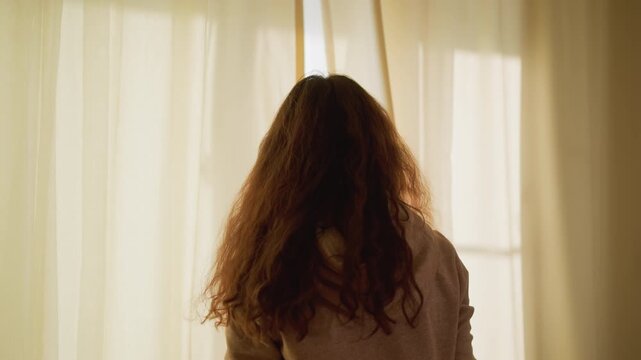 Woman opens white curtains letting in soft morning sunlight. Her back and hair are visible as woman pulls sheer fabric aside revealing warm glow from outside. scene is calm with diffused