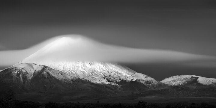 Black and White image of snow-capped Mt Ngauruhoe partially draped in a dramatic lenticular cloud. Tongariro National Park. New Zealand.