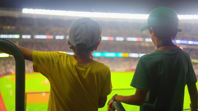 Two young boys watching a baseball game at night from the stadium stands, enjoying the exciting competition and the bright lights of the professional sports arena