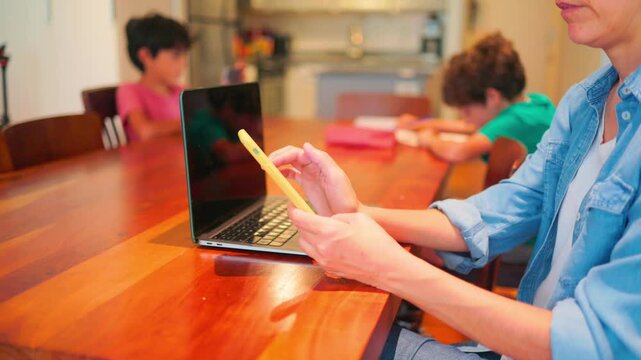 Mother working from home with a laptop and smartphone while two children are homeschooling in the background, a new normal concept during a global pandemic