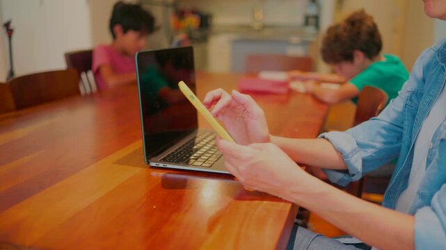 Mother multitasking using a smartphone and laptop for work while her children are doing homework at the dining room table, illustrating the challenges of modern family life