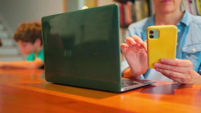 Woman working from home using her smartphone and laptop while her young son is studying in the background, representing remote work, multitasking and family life