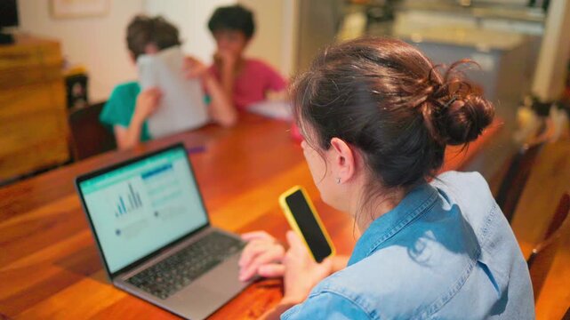 Mother juggling work from home and homeschooling her children, multitasking with a laptop and smartphone while kids study in the background at the dining room table