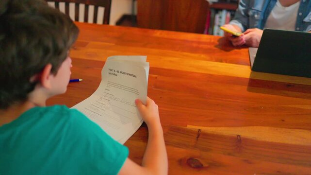 Focused elementary student concentrating and carefully reading his school test paper while his parent checks her smartphone sitting across the wooden table at home