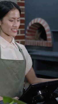 Vertical video: Taking customer order, barista in green apron using POS and marking cup at cafe