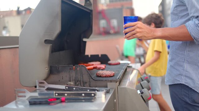 Family enjoying barbecue party on rooftop patio, father grilling hamburgers and holding blue cup with friends and children enjoying food and drink in the background