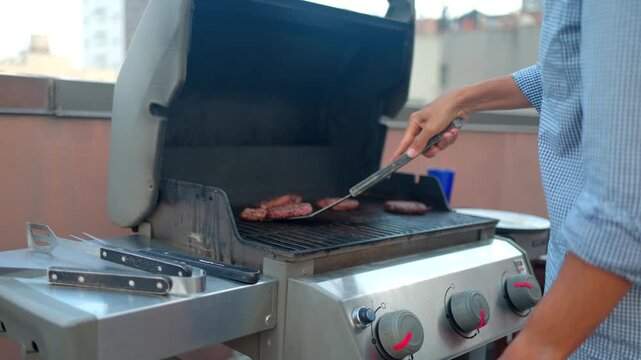 Man grilling beef hamburgers on a rooftop gas barbecue during a summer party, flipping meat patties with a spatula while enjoying an outdoor cookout in the city