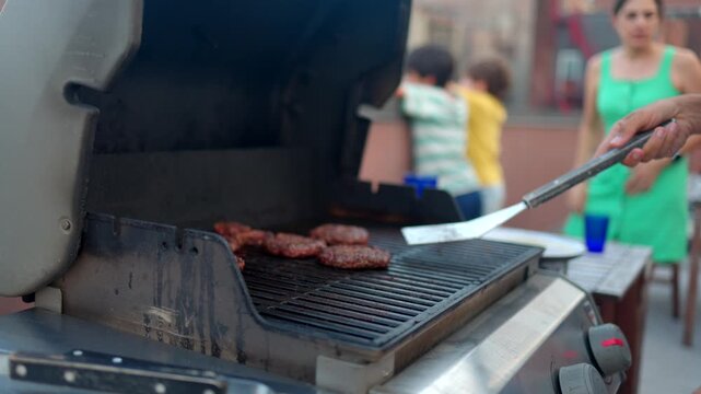 Close-up of a man's hands grilling beef hamburger patties on an outdoor barbecue during a summer family party, using a spatula to flip the delicious meat