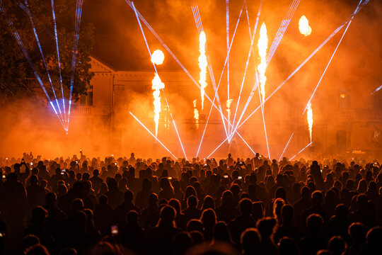 Dramatic pyrotechnic fire pillars and orange laser lights illuminating large crowd.