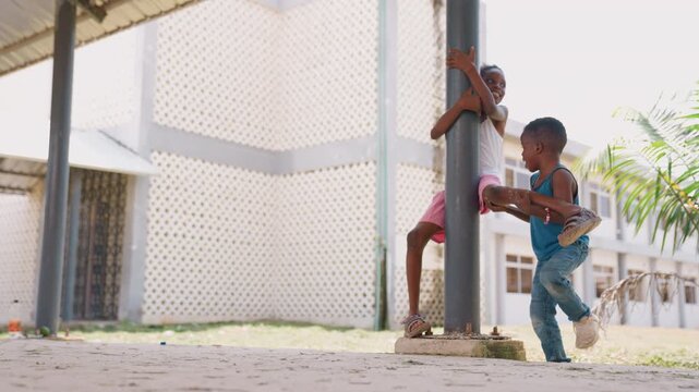 Children engage in playful interactions outdoors one child climbs pole while another approaches. scene features bright daylight and exterior building with lattice walls suggesting casual