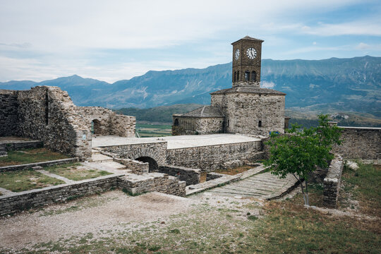 Historic clock tower standing on stone fortress grounds against a scenic mountain backdrop in Gjirokaster Castle, Albania.
