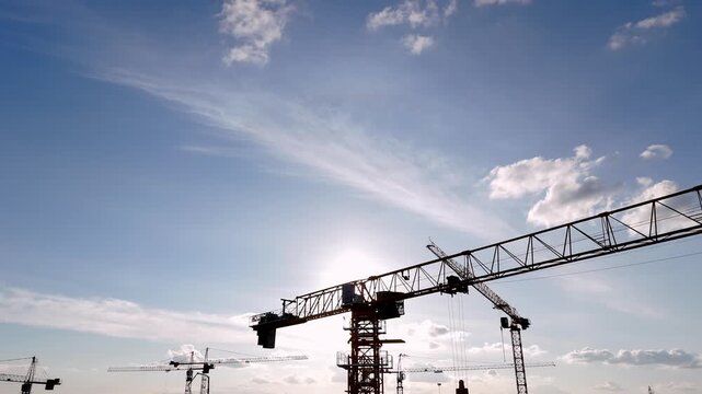 Silhouetted cranes rise against a bright blue sky. Sunlight glints off metal arms and distant structures. Workers move quietly beneath towering machinery. Construction buzzes beneath wide open skies
