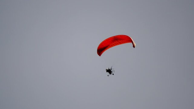 Red parachute soars high against pale sky. Parachutist descends calmly, relaxed and free. Perfect for adventure, freedom, or sky sports visuals. No animals or plants present