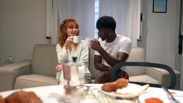 Young mixed race couple enjoying a morning conversation while sitting on the couch and drinking coffee together at home, with croissants and a moka pot visible on the table in the foreground