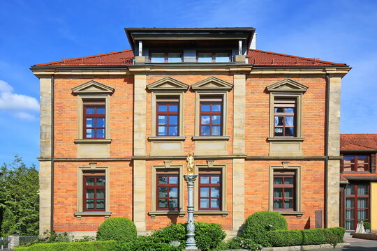 Prachtvolle Klinkerfassade des historischen Geb&auml;udes in Auerbach in der Oberpfalz mit einer goldenen Mariens&auml;ule im Vordergrund vor einem strahlend blauen Himmel.