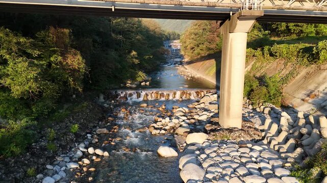 The underside of a large road bridge and concrete pillar frame multiple river cascades running between rocky banks.