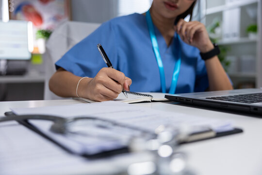 Medical professional writing notes at desk beside laptop and medical records, focused on documentation in clinical office